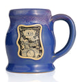 The front of the Bones Coffee Company Blueberry Blast Cake hand thrown mug. The golden medallion has the art of the Blueberry Blast Cake coffee on it. The mug is sky blue finish dipped in white and lilac glaze.